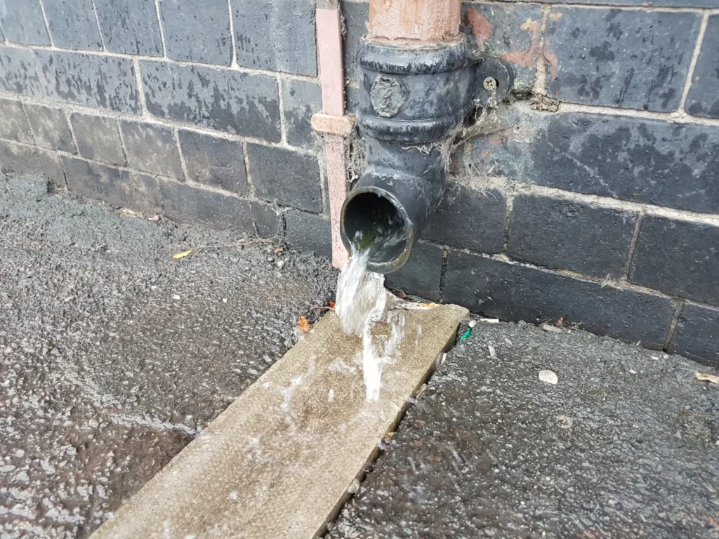 Downspout discharging rainwater onto paving at the base of a stone wall