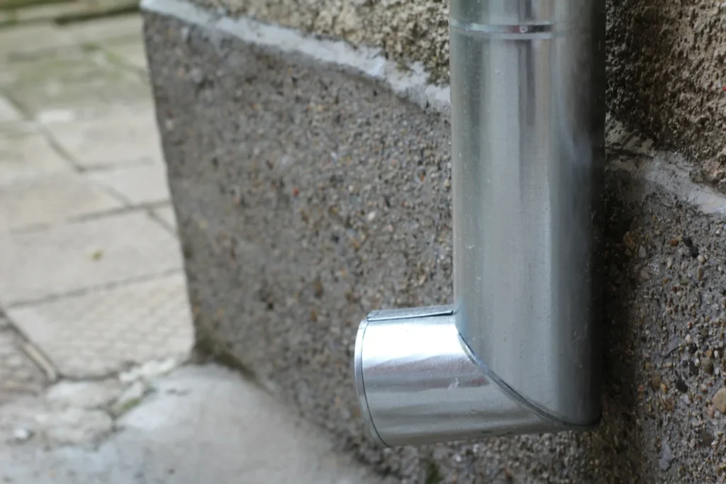 Metal downspout elbow at the base of a wall where roof water leaves the house